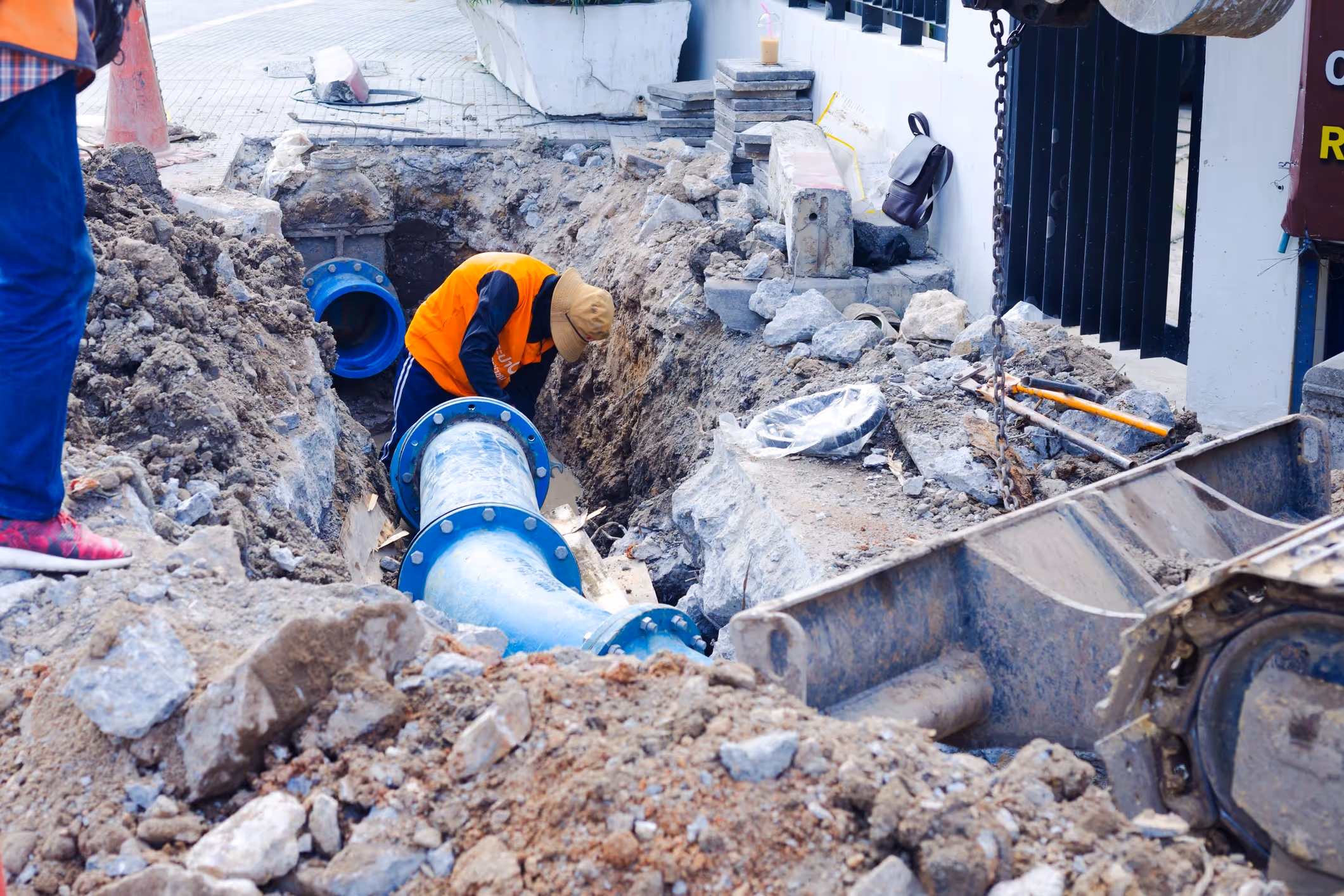 Worker in an orange vest and hat installs large blue pipes in a trench. 