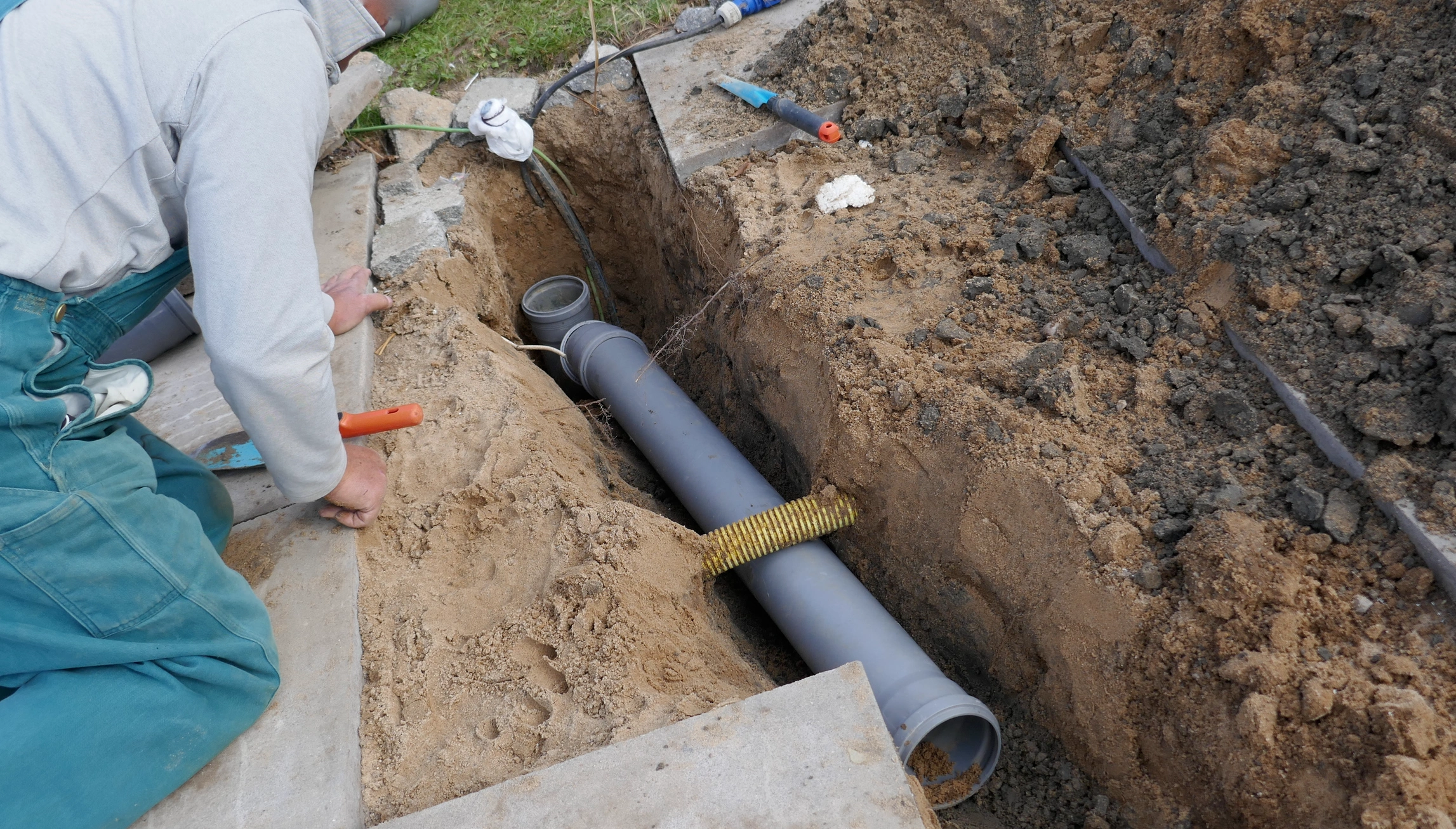 A worker in green overalls and gloves installs a gray drainage pipe in a trench.