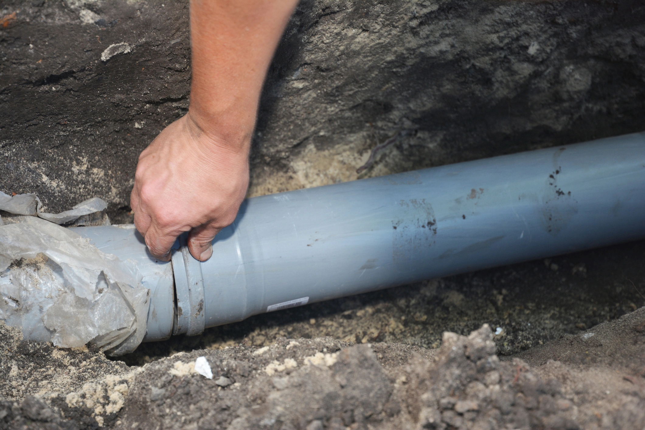 A person's hand grips a blue PVC pipe in a trench surrounded by soil. 