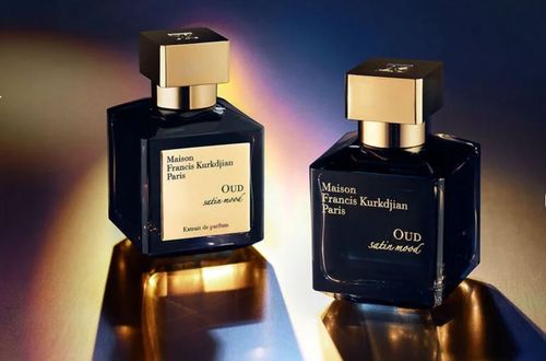 Two bottles of Maison Francis Kurkdjian Oud Satin Mood perfume with gold caps, displayed under dramatic lighting on a reflective surface.