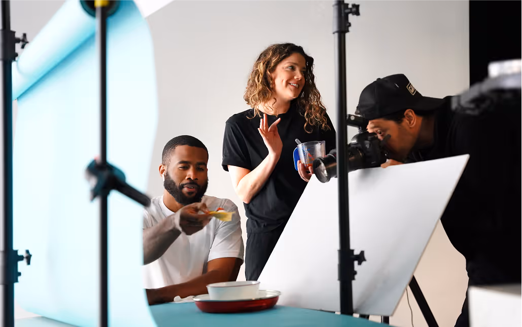 A man holds a piece of food near a photographer's camera during a studio photo shoot