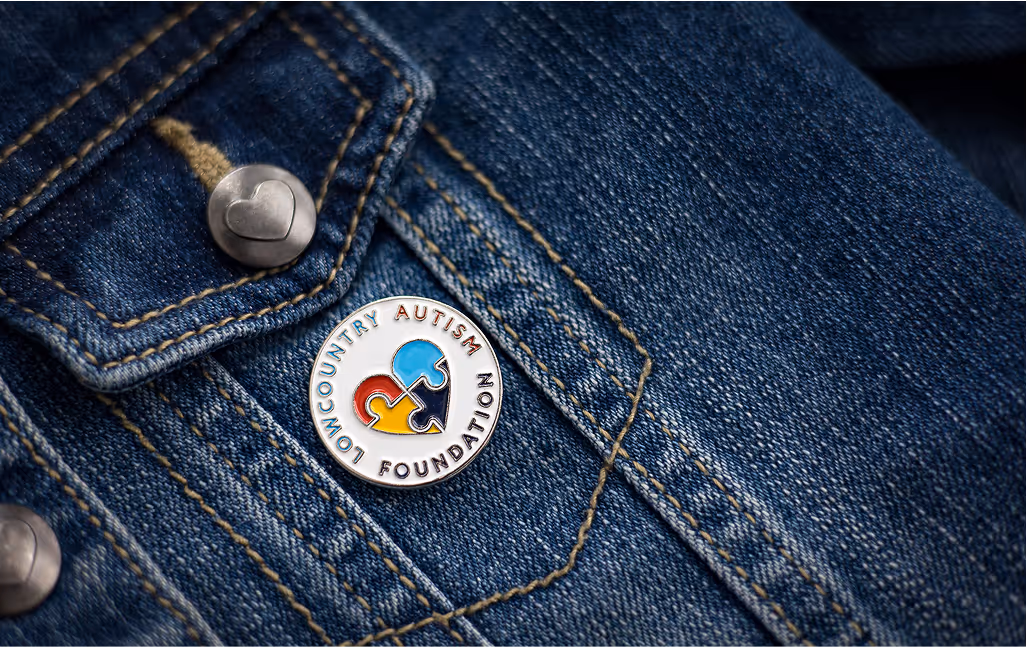 Close up view of a circular Low Country Autism Foundation pin with a white background, displayed on a denim jacket
