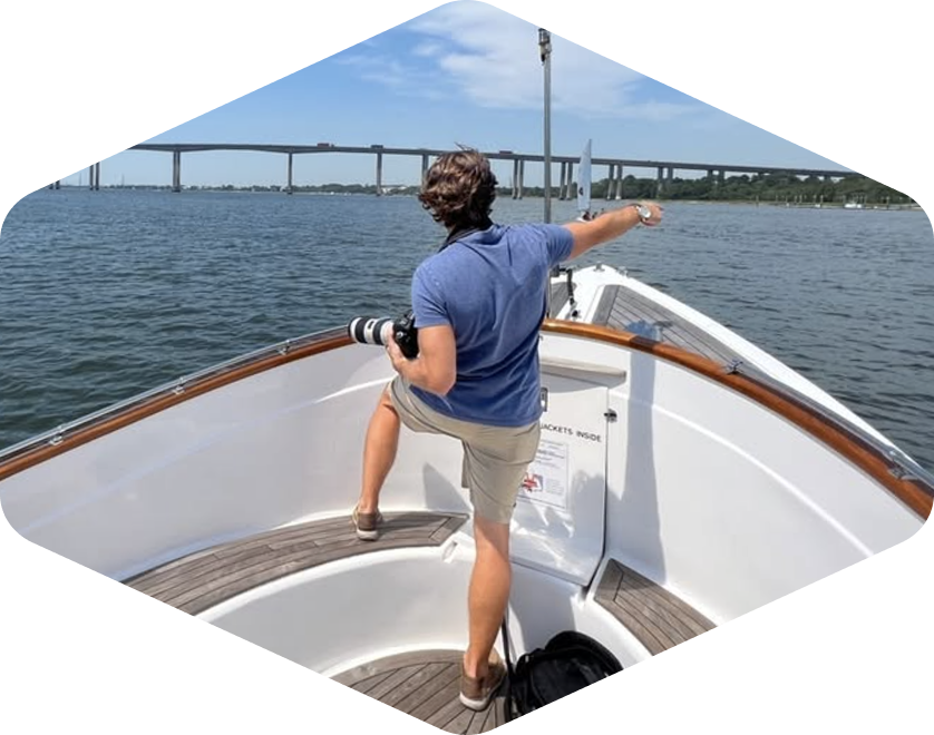 Male photographer on a boat pointing toward a scenic bridge in Charleston, South Carolina, with DSLR camera in hand and clear blue skies overhead.