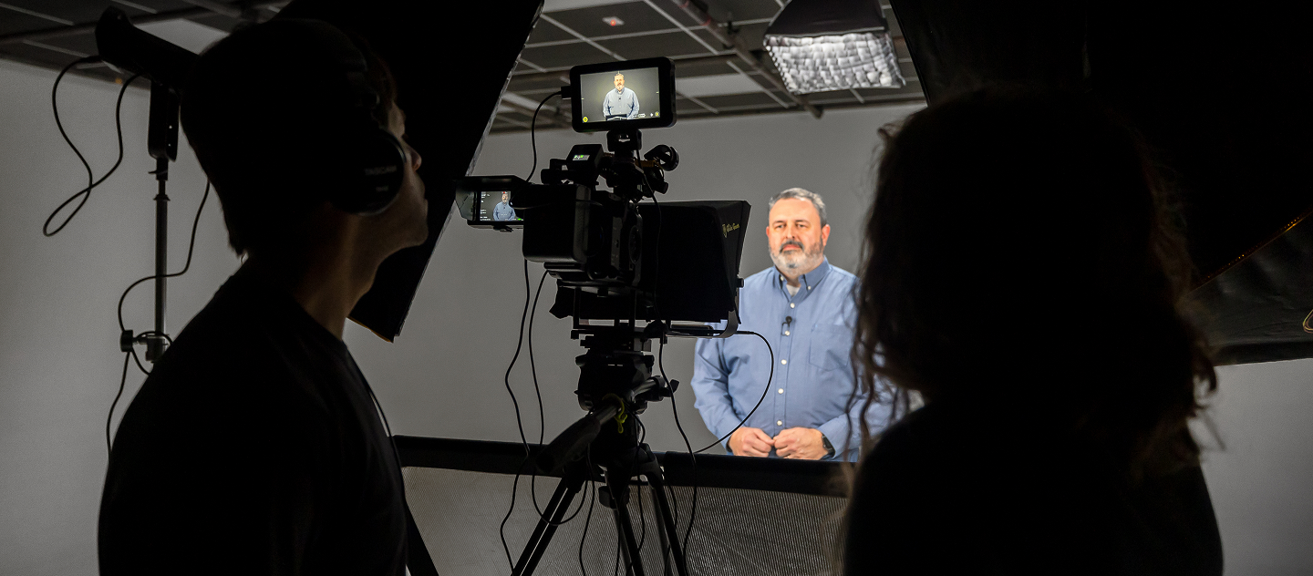 Professional video shoot of man speaking on camera in studio interview setup with lighting and crew in the background.