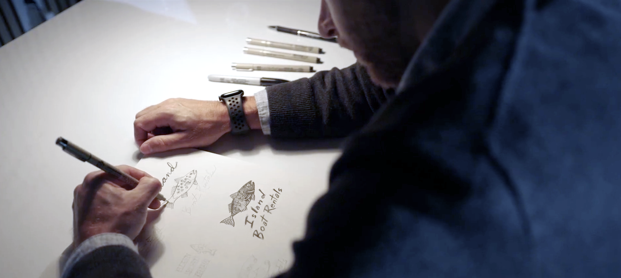 Designer sketching a fish-themed logo for Island Boat Rentals by hand on a white desk with multiple fine-tip pens