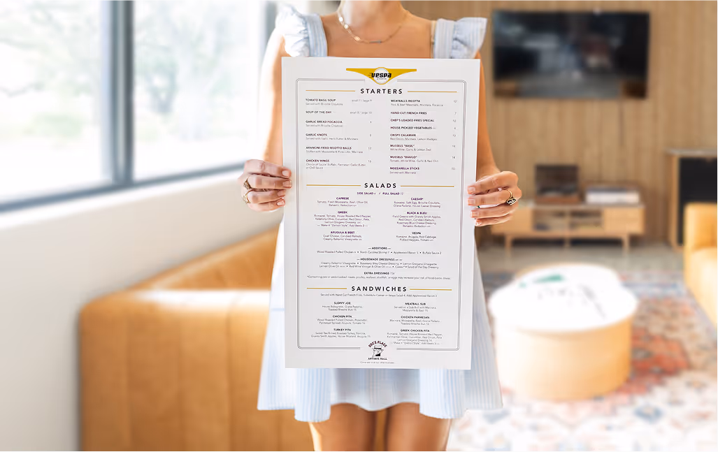Woman holding full-size printed Vespa Pizzeria menu showing starters, salads, and sandwiches, with interior lounge visible in background