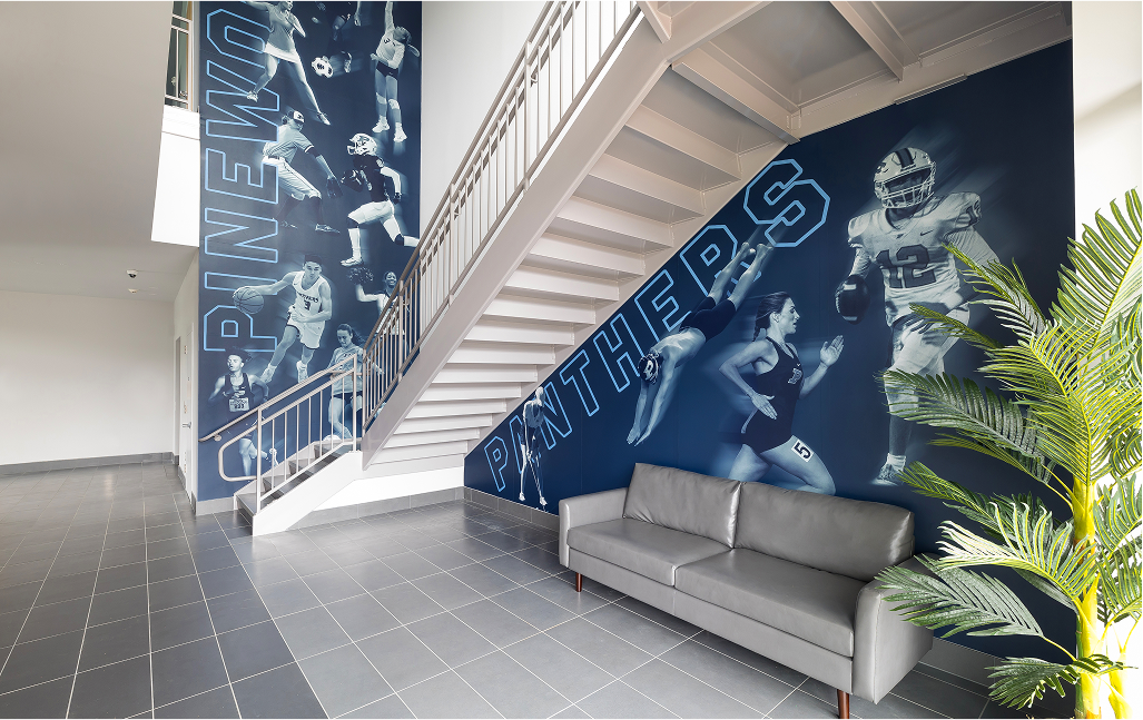 Interior stairwell mural featuring Pinewood Panthers athletes in action, with bold typography on deep blue walls
