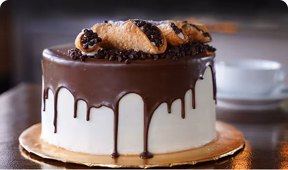 Chocolate-drizzled cannoli cake with white frosting and mini chocolate chips, served on a gold cake board at Carmella’s Cafe & Dessert Bar in Charleston.