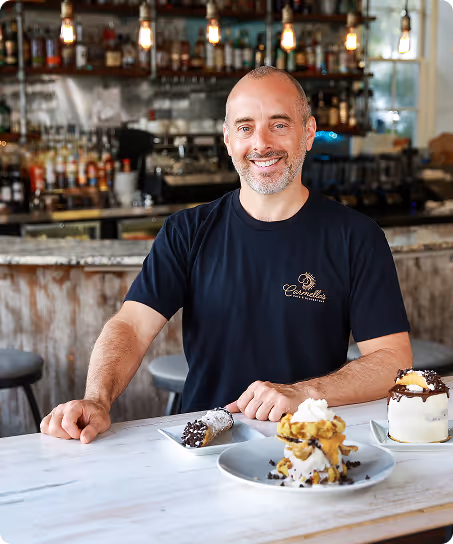 Owner of Carmella's Bakery displaying various deserts available at his shop.