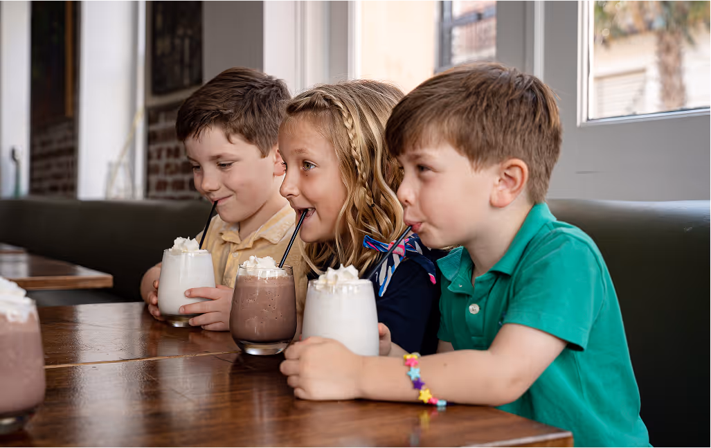 Three smiling children at Carmella’s Cafe enjoying milkshakes with whipped cream at a wooden booth.