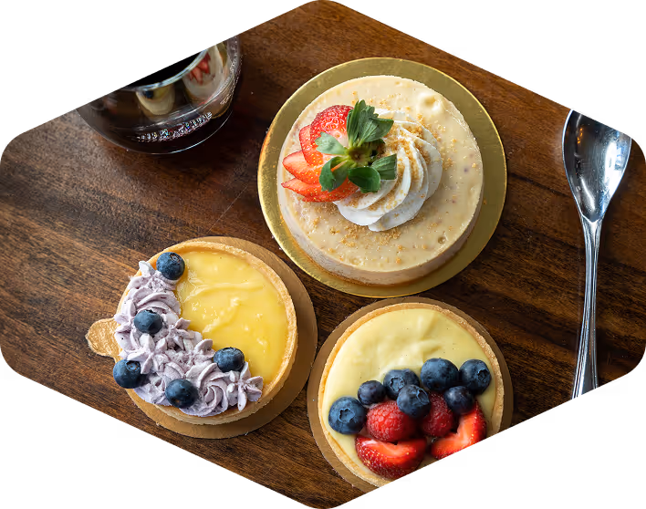 Overhead view of three artisan fruit tarts at Carmella’s Café, including lemon curd with blueberries, vanilla custard with strawberries and blueberries, and cheesecake with whipped cream and fresh strawberries, served on a wooden table.