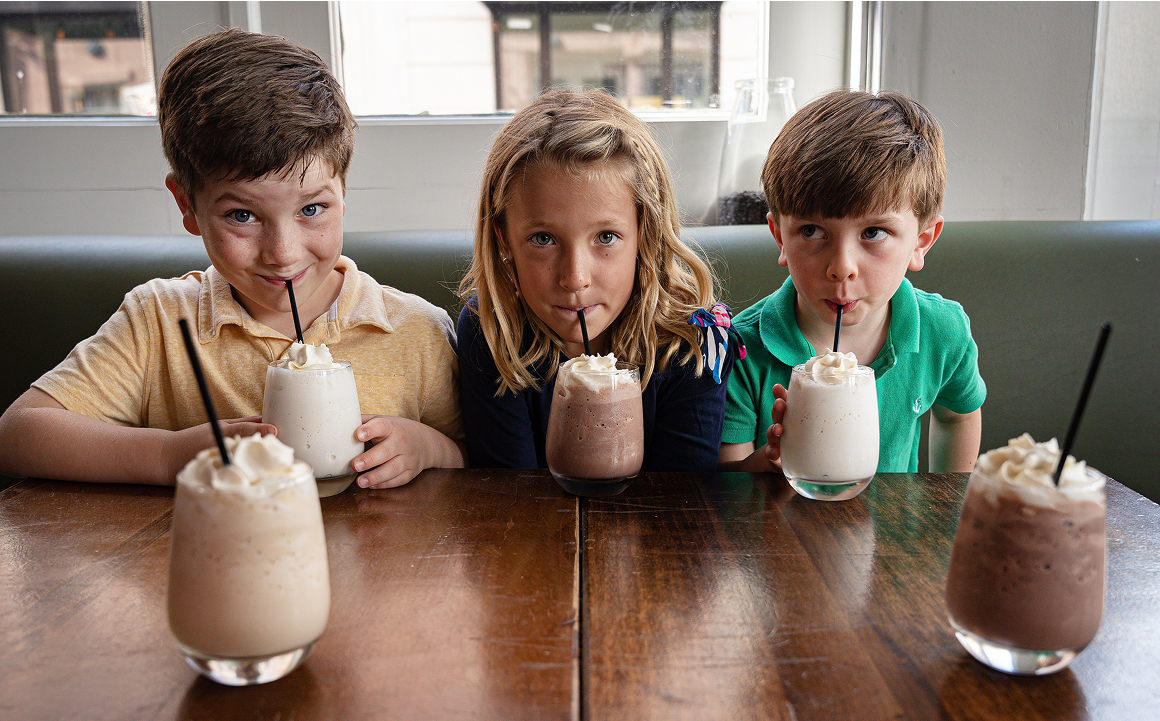 Three smiling children at Carmella’s Cafe enjoying milkshakes while sitting in a wooden booth.