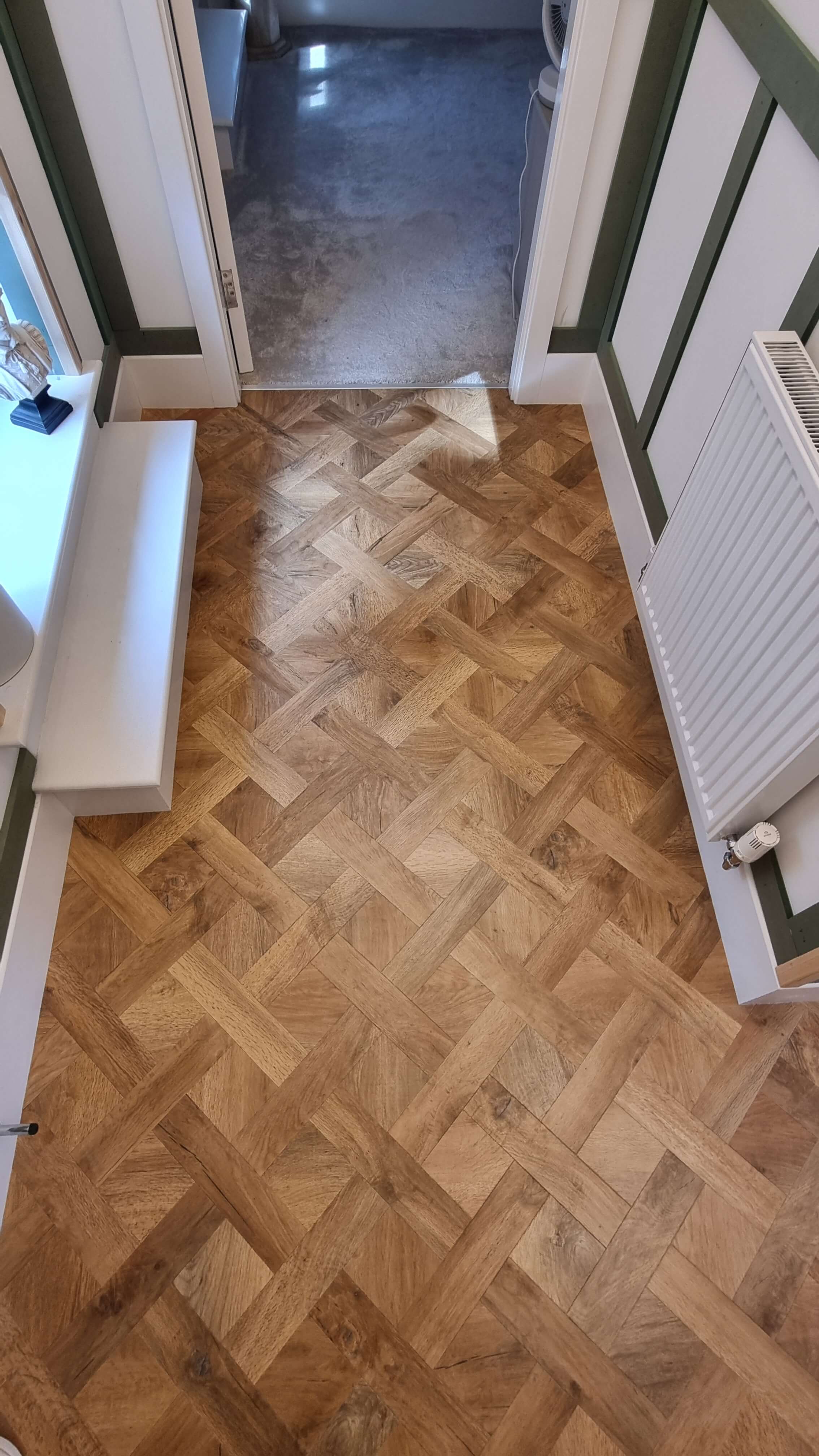 Wooden parquet floor with a geometric pattern in a hallway leading to a carpeted room, white radiator on the right, a small white bench on the left, and white paneled walls with green trim.