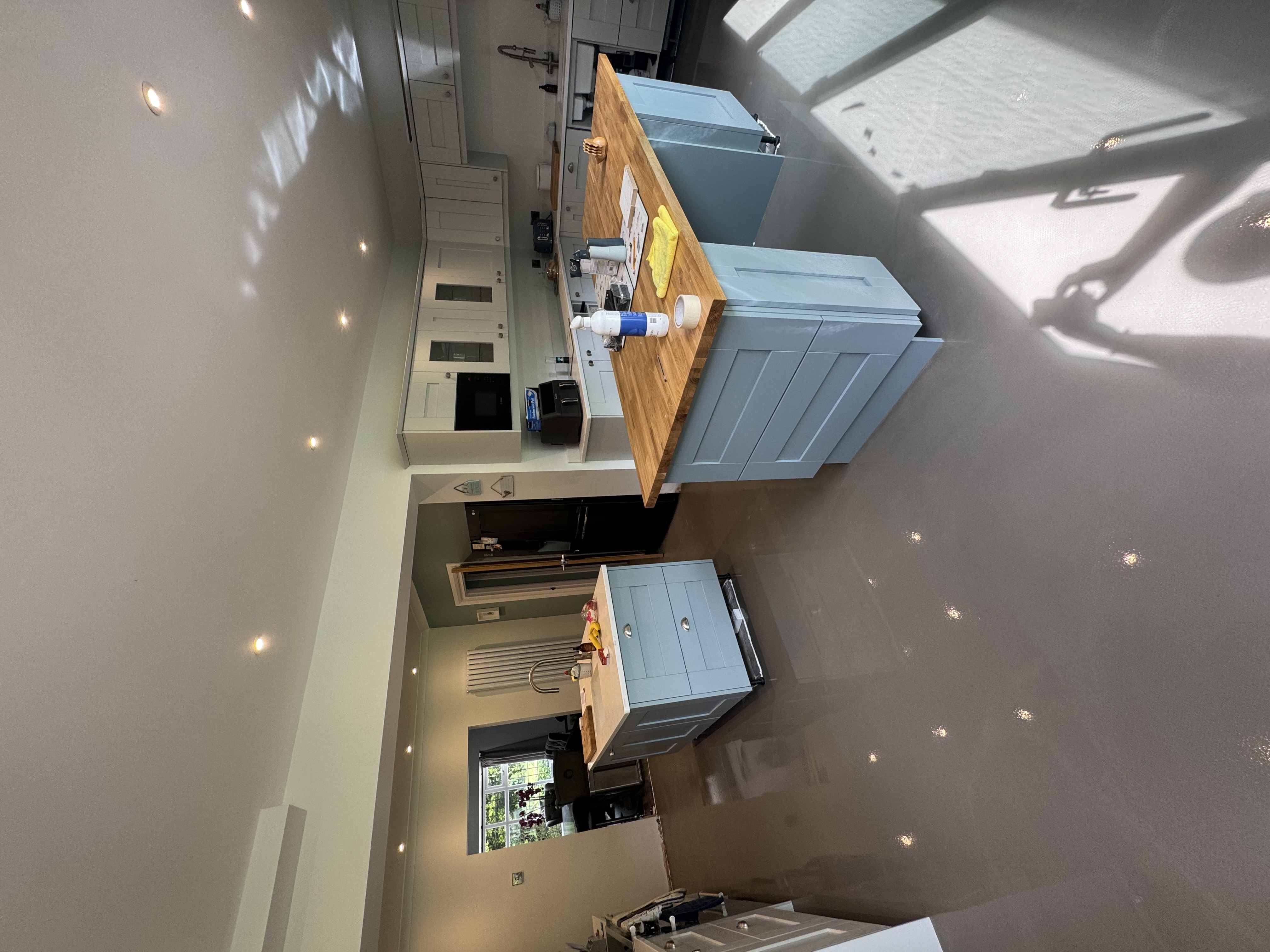 Spacious modern kitchen with two light blue islands, one with a wood countertop and cleaning supplies on top, glossy gray floor, white upper cabinets, black refrigerator, and stainless steel faucet, lit by recessed ceiling lights and natural light from large windows.