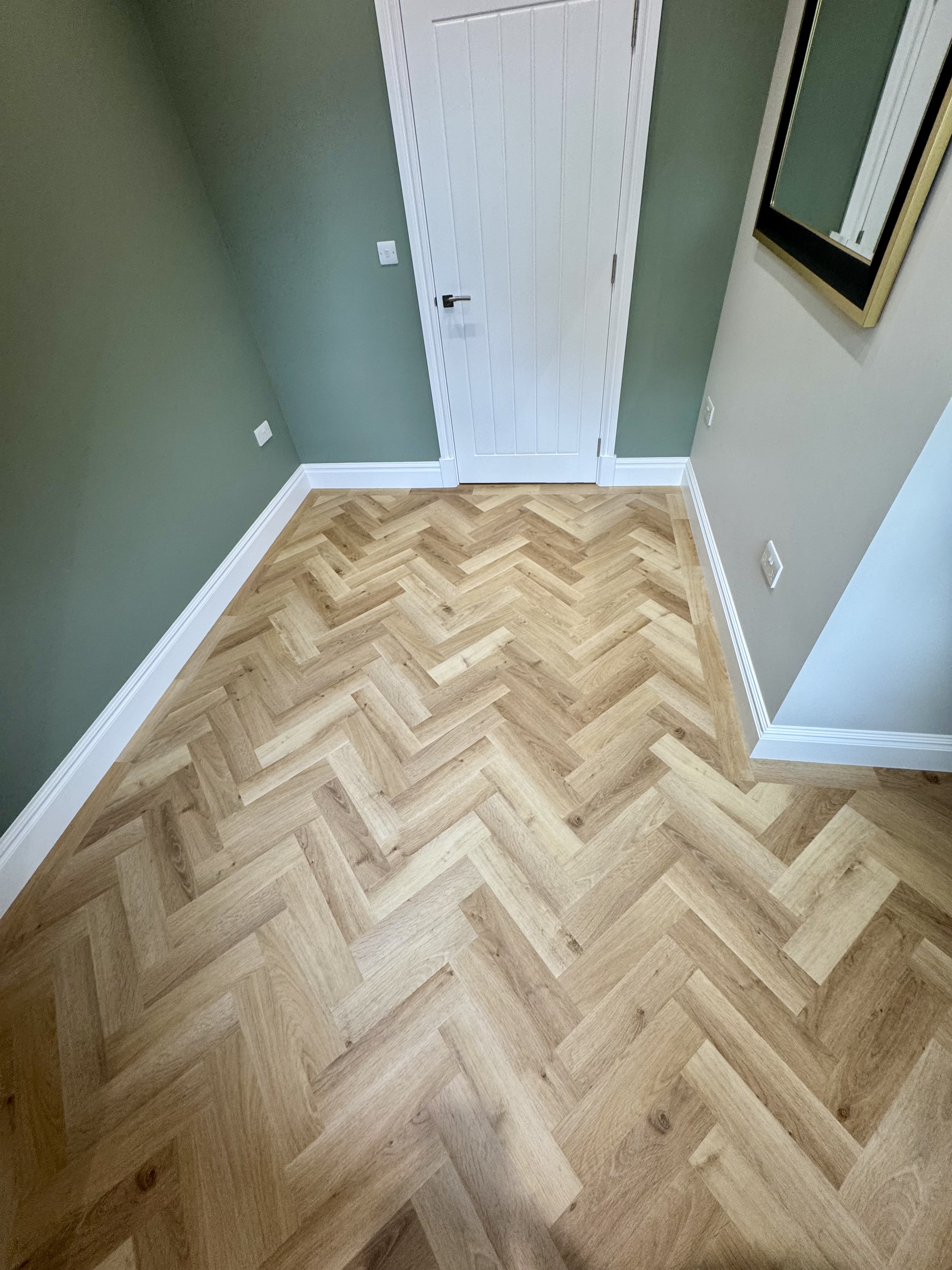 Entryway with chevron-patterned light wood floor, green-painted walls on the left and center, white door with vertical panels, and a beige wall on the right with a rectangular mirror.