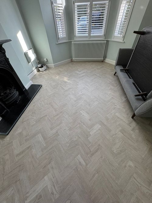 Room with light wood herringbone patterned floor, light blue walls, white radiator under bay window with white shutters, black fireplace on left, and gray sofa on right with its seat cushion flipped up.