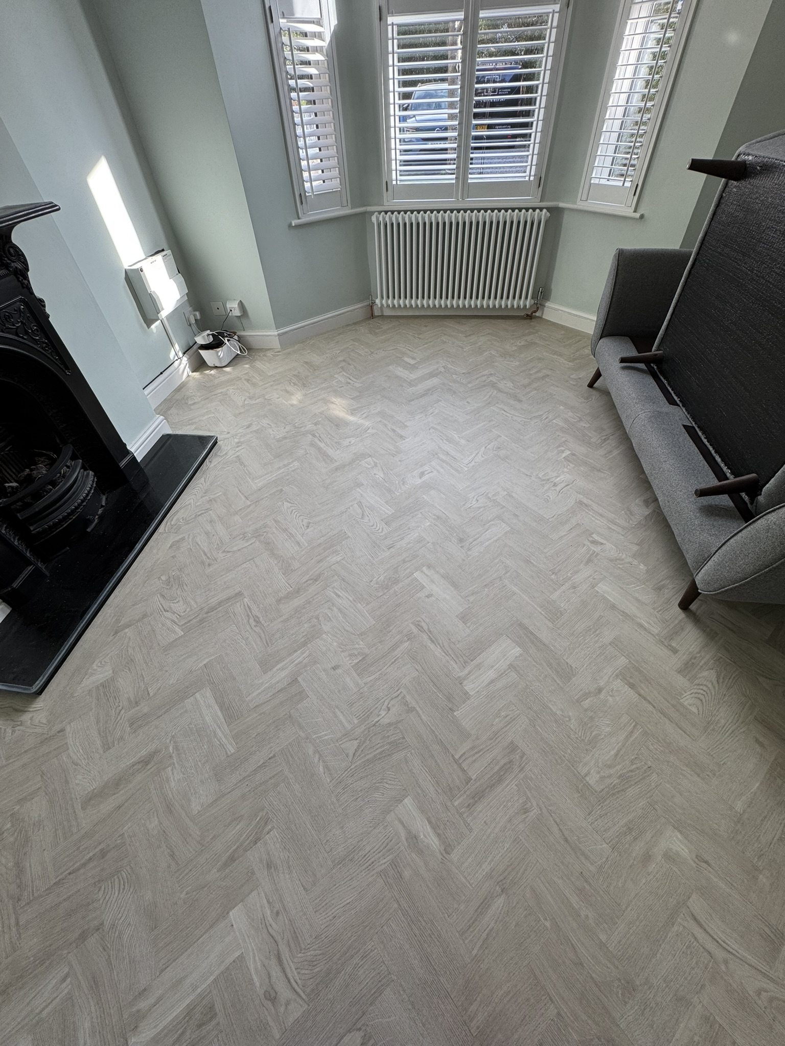 Room with light wood herringbone patterned floor, light blue walls, white radiator under bay window with white shutters, black fireplace on left, and gray sofa on right with its seat cushion flipped up.