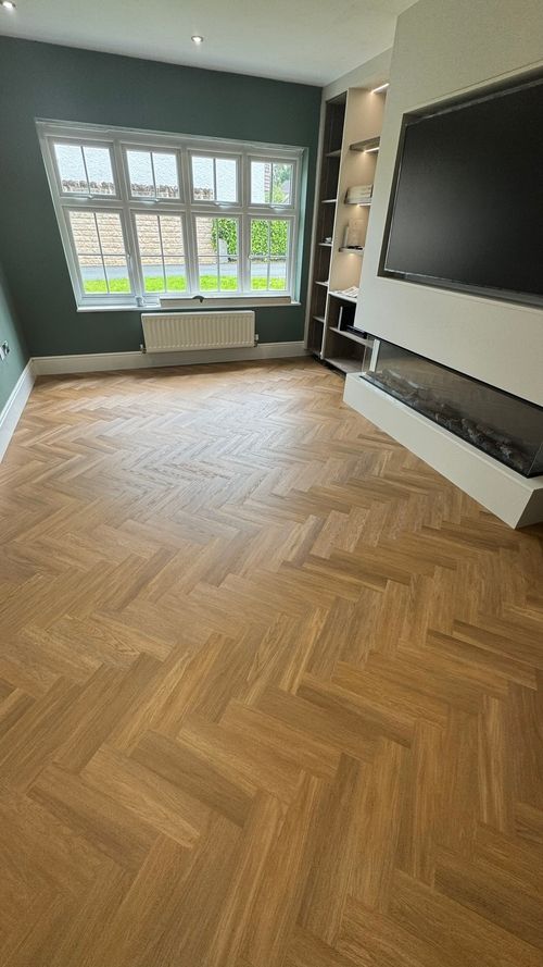 Modern living room with herringbone-patterned wooden floor, green accent wall with large white-framed window, white radiator below the window, built-in shelves with recessed lights, wall-mounted flat-screen TV, and a sleek glass-front fireplace.
