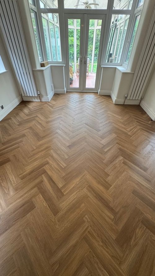 Empty room with light brown herringbone wood flooring, cream walls, tall white vertical radiators on each side, and glass double doors leading to a patio with greenery outside.