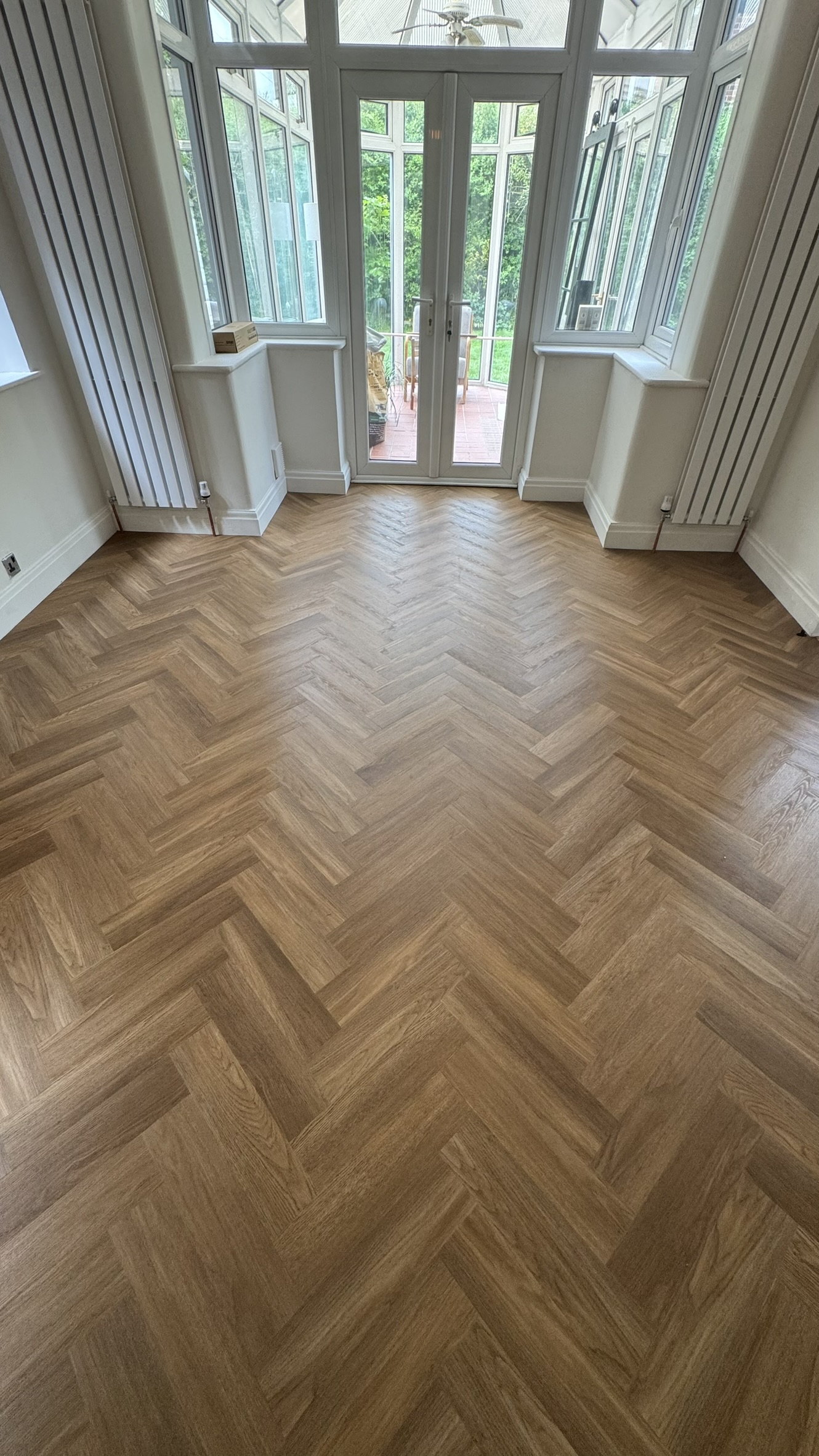 Empty room with light brown herringbone wood flooring, cream walls, tall white vertical radiators on each side, and glass double doors leading to a patio with greenery outside.