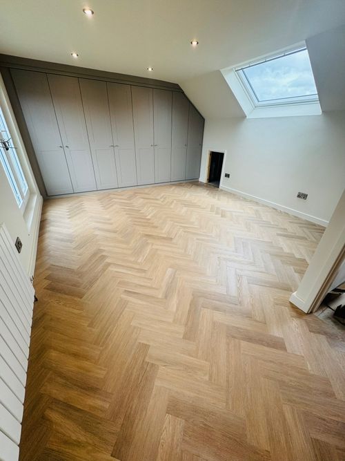 Empty attic room with light wood herringbone-patterned floor, gray paneled built-in cabinets along one wall, a large skylight on the sloped ceiling, white walls, and a small low door near the cabinets.