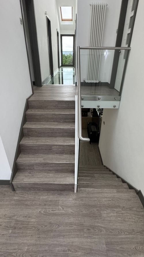 Indoor staircase with light wood flooring and a metal handrail with glass paneling, leading up to a hallway with a skylight, white walls, and a radiator, ending in a glass door showing trees outside.