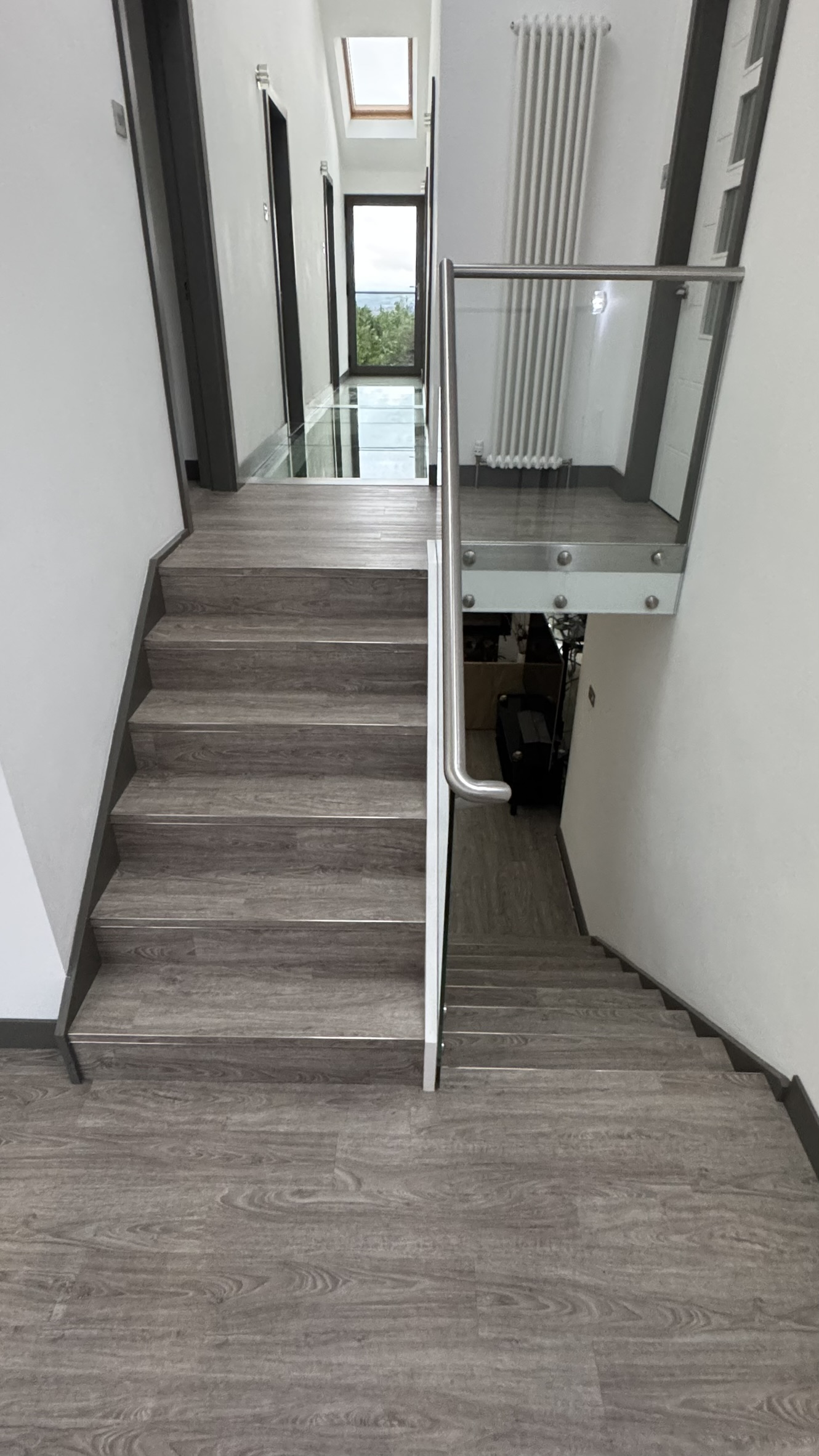 Indoor staircase with light wood flooring and a metal handrail with glass paneling, leading up to a hallway with a skylight, white walls, and a radiator, ending in a glass door showing trees outside.