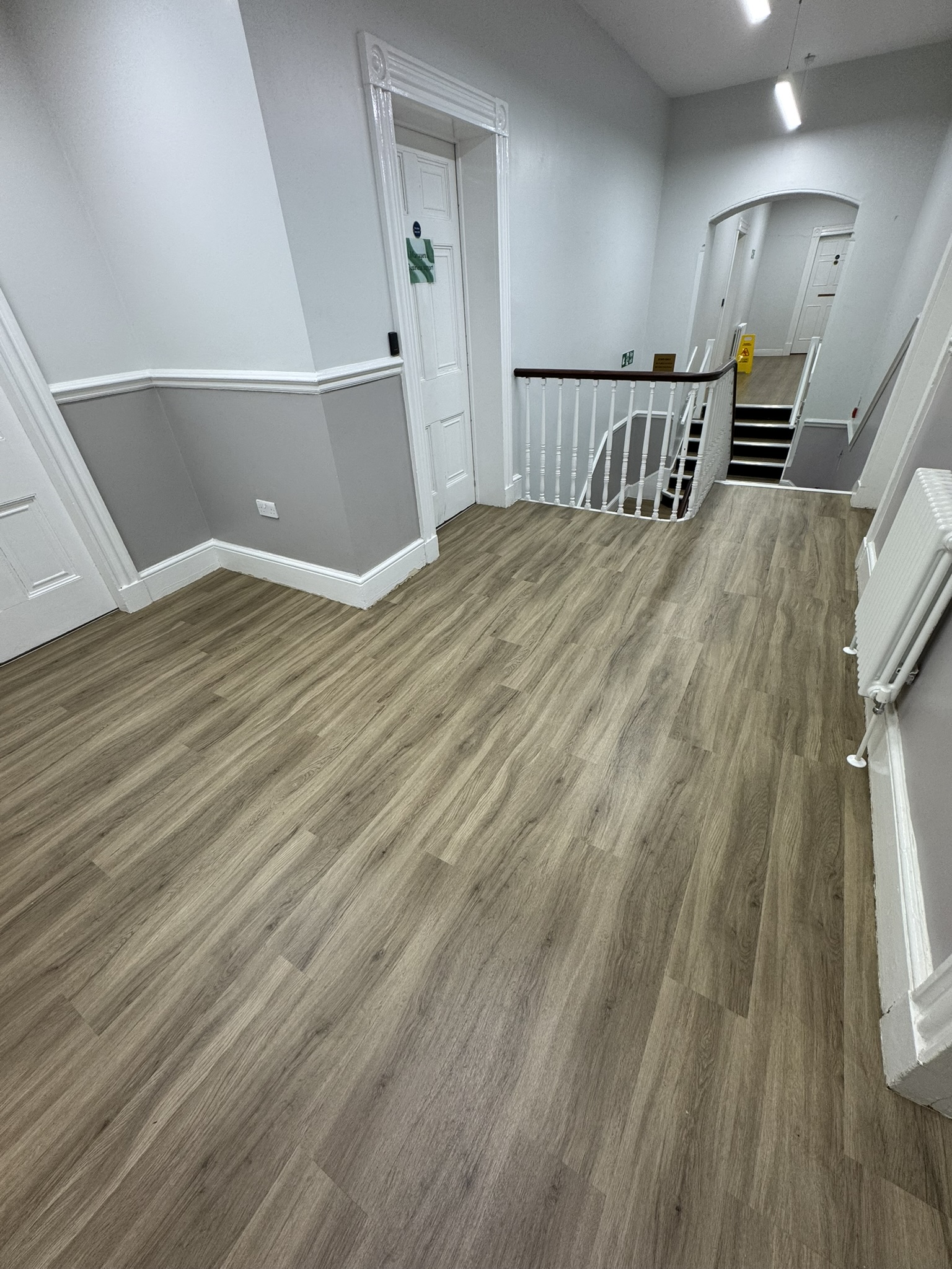 Interior hallway with light wood flooring, white and gray walls, white doors, white banister around staircase, a radiator on the right wall, and fluorescent ceiling lights.
