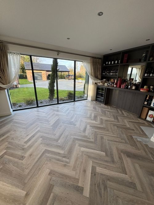 Modern room with large floor-to-ceiling windows showing a green lawn and brick garage outside, light herringbone wood flooring, beige curtains partially tied back, and a dark wood built-in bar cabinet stocked with various bottles and a wine cooler.