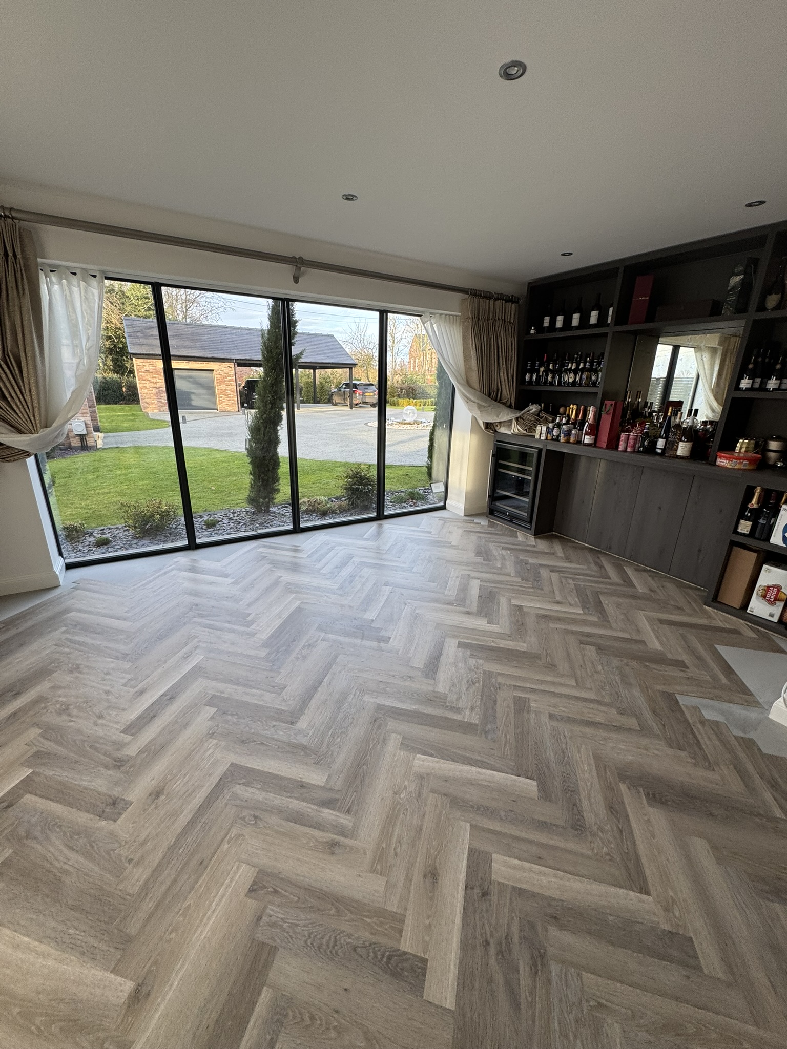 Modern room with large floor-to-ceiling windows showing a green lawn and brick garage outside, light herringbone wood flooring, beige curtains partially tied back, and a dark wood built-in bar cabinet stocked with various bottles and a wine cooler.
