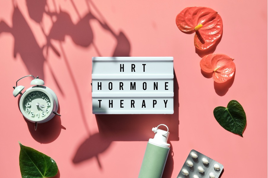 Flat lay of hormone replacement therapy concept: lightbox reading “HRT Hormone Therapy,” alarm clock, pills, and tropical leaves on a pink background.
