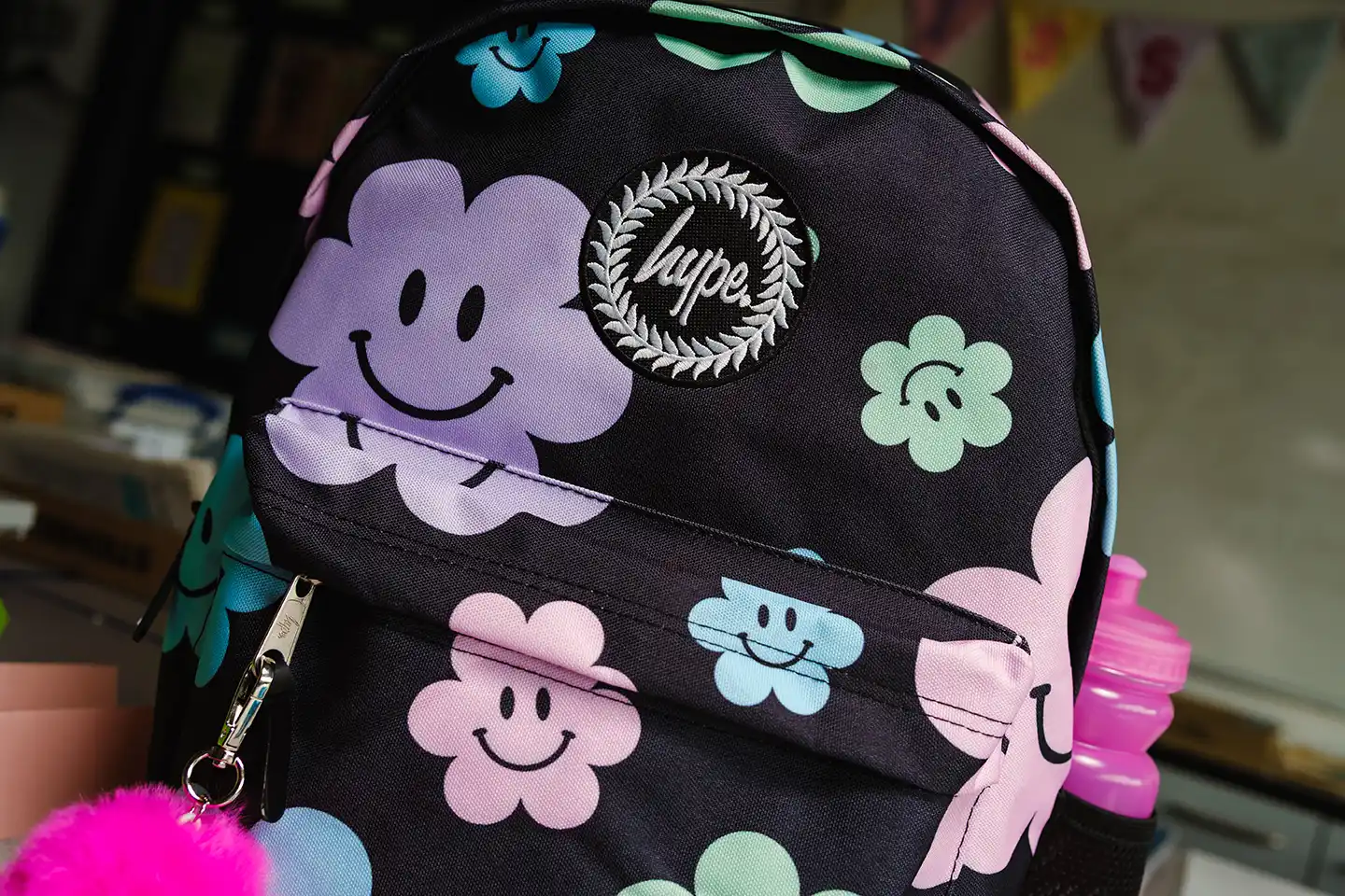 Black backpack with colorful smiling flower patterns, a pink fluffy keychain, and a pink water bottle in side pocket.