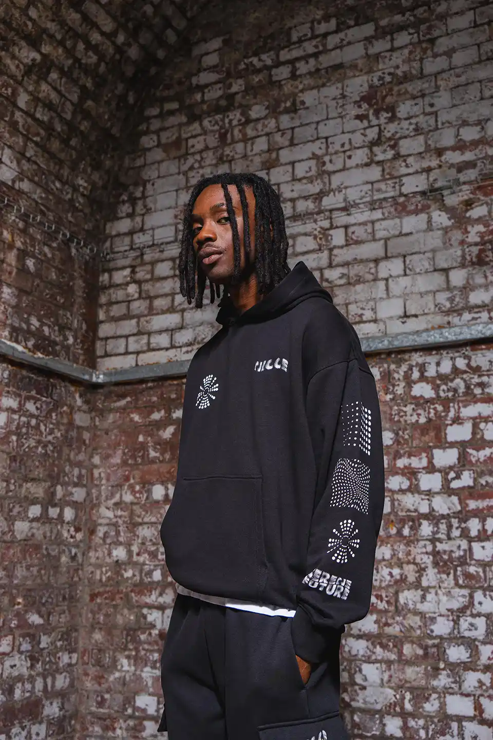 Man wearing a black hoodie with white abstract designs and text standing against a weathered brick wall.