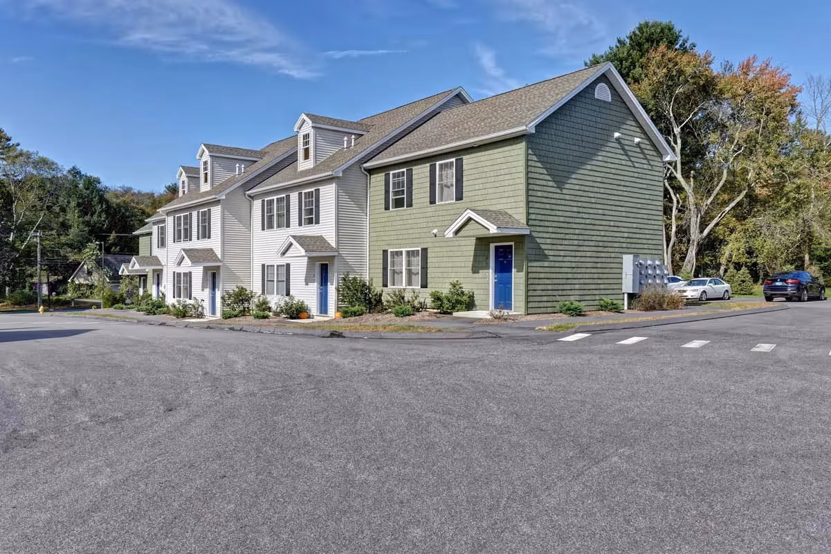 Exterior view of light-colored townhome-style apartment buildings with blue doors and black shutters, featuring dormer windows, at Meadowbrook Gardens in Mansfield Center, CT.