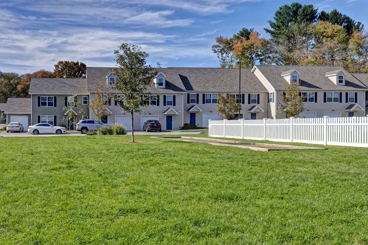 Exterior view of light-colored townhome apartment buildings with dormer windows and a white picket fence, at Meadowbrook Gardens in Mansfield Center, CT.