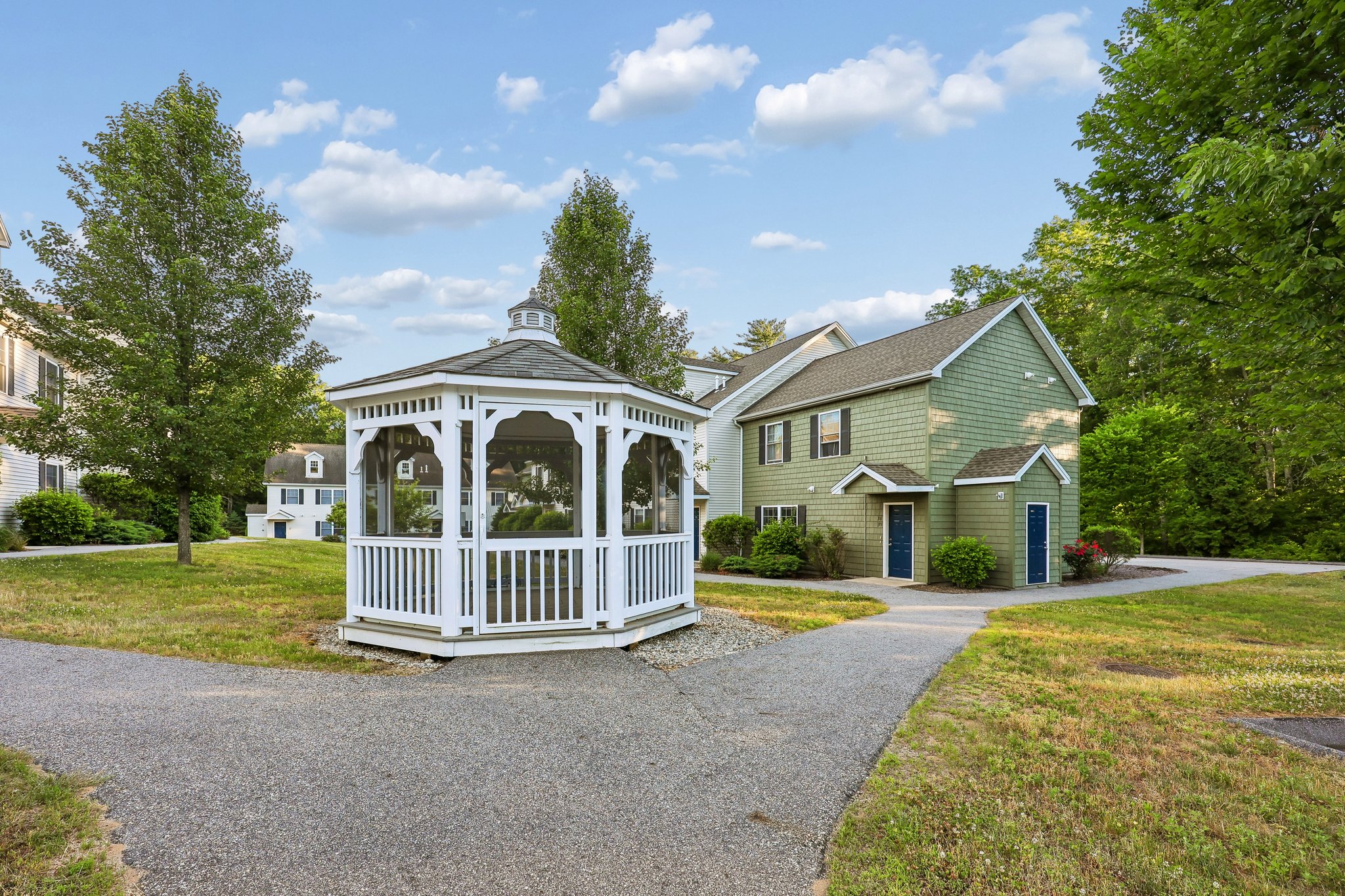 White Gazebo and outdoor view of Meadowbrook Gardens apartment community in Mansfield Center, CT, showing rows of townhomes nestled in a green, wooded landscape.
