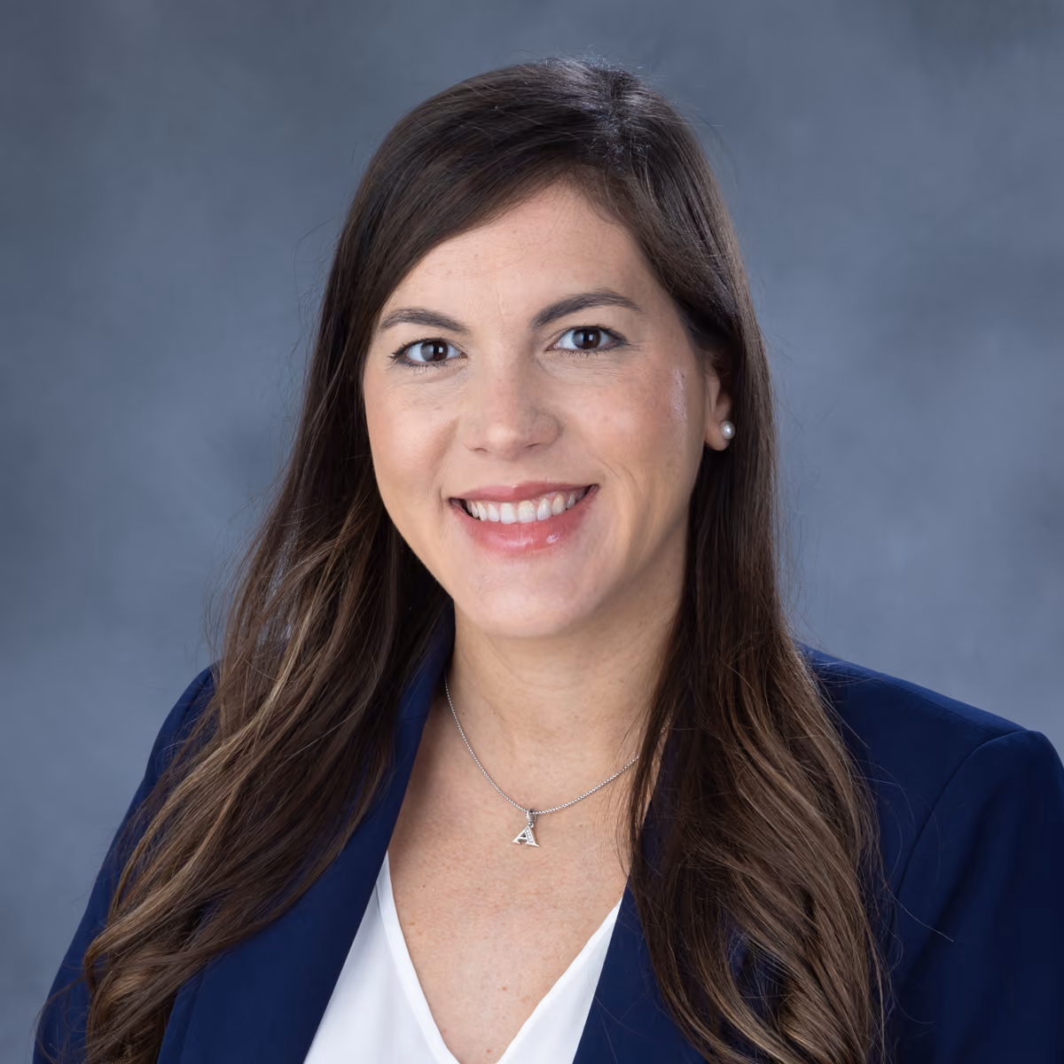 Smiling woman with long brown hair wearing a navy blazer and white top against gray background.