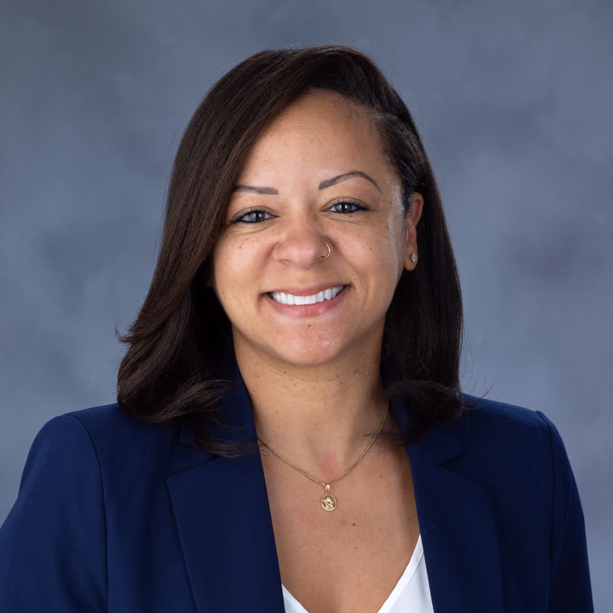 Smiling woman with dark hair wearing a navy blazer, white top, gold necklace, and nose ring on gray background.
