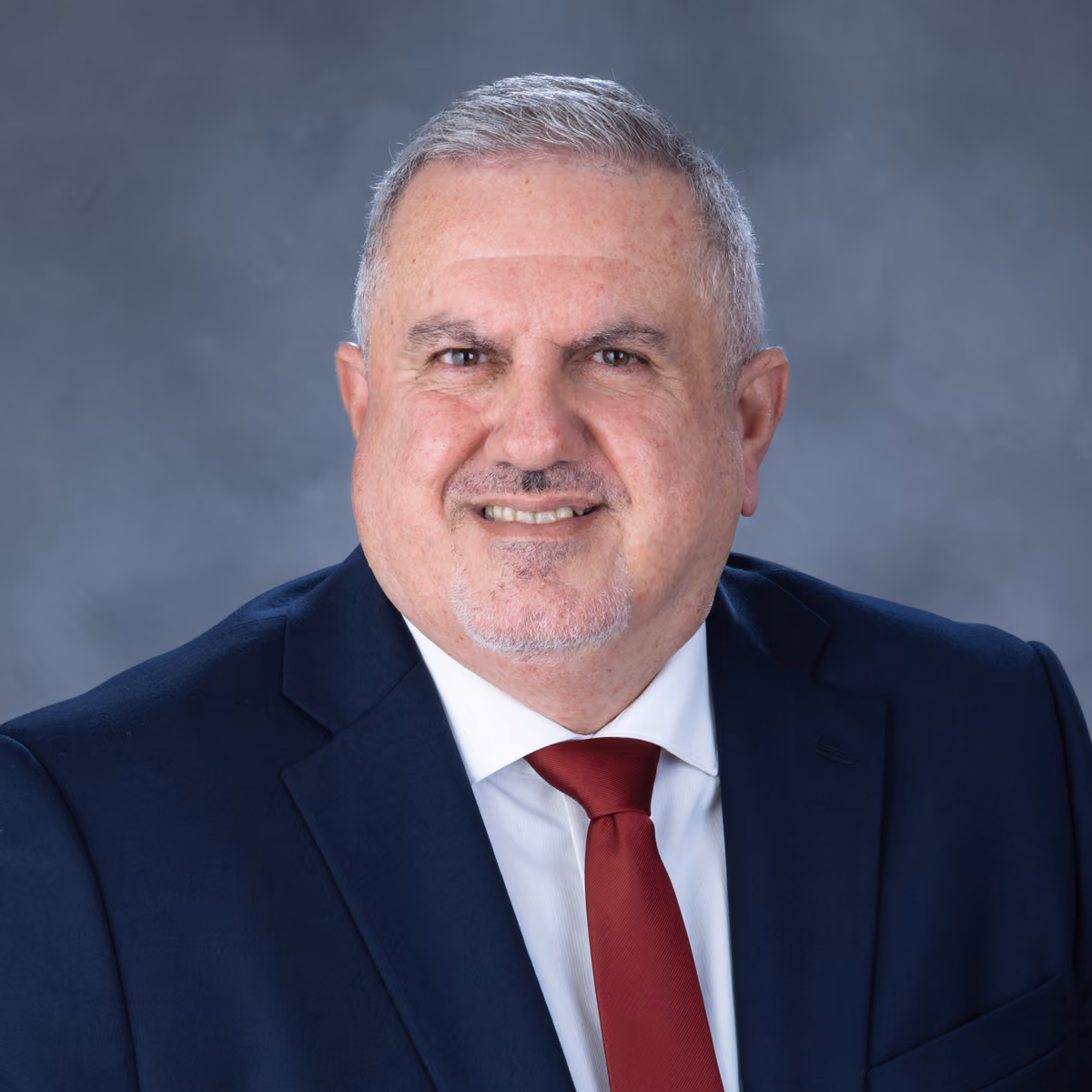 Smiling middle-aged man with gray hair wearing a dark suit, white shirt, and red tie against gray backdrop.