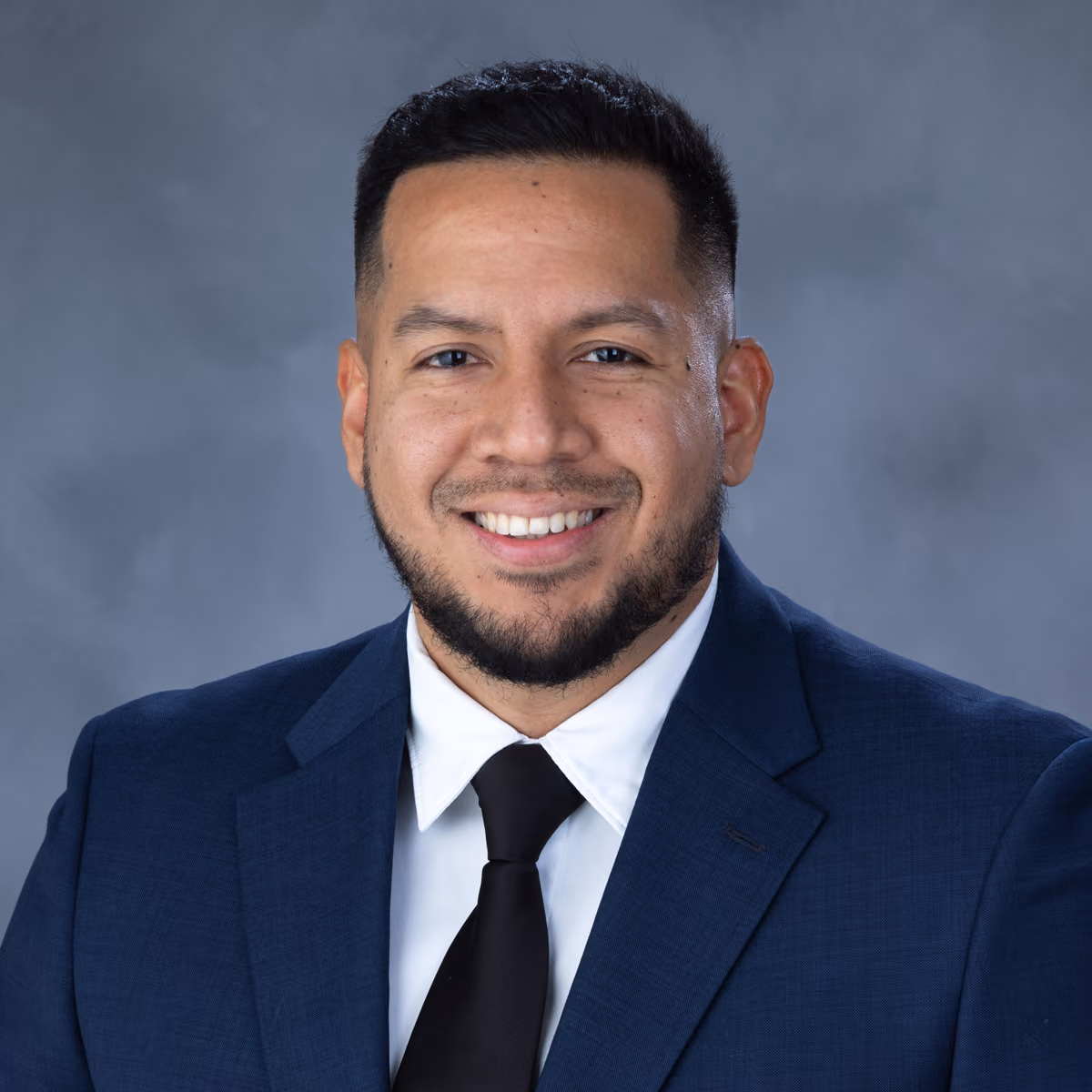 Smiling man with short hair, beard, blue suit, white shirt, and black tie against a gray background.