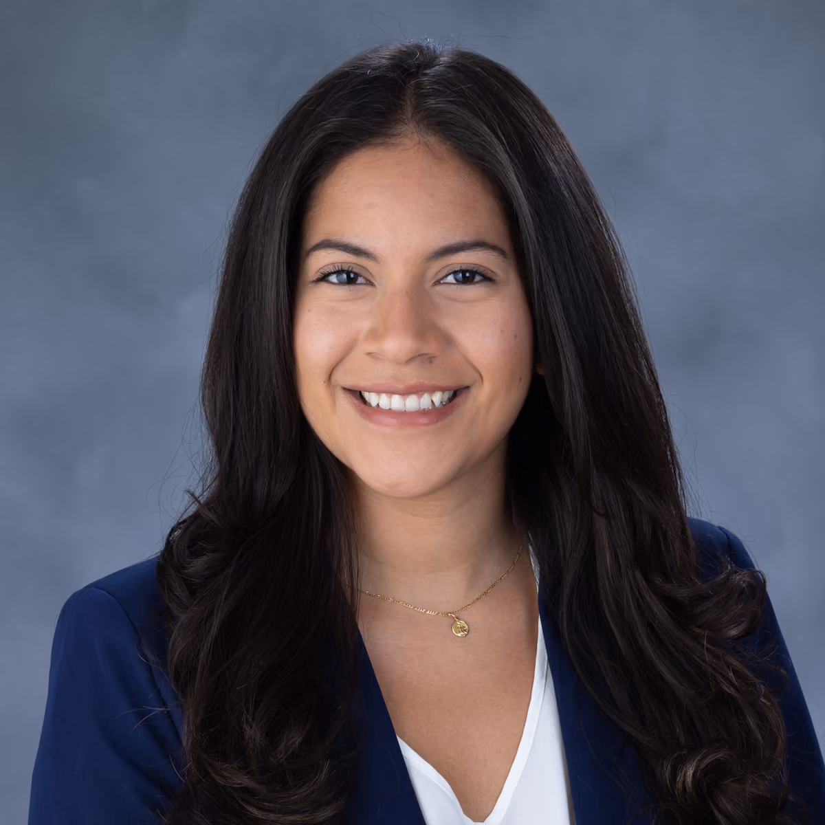 Smiling woman with long dark hair wearing a blue blazer and white top against a gray background.