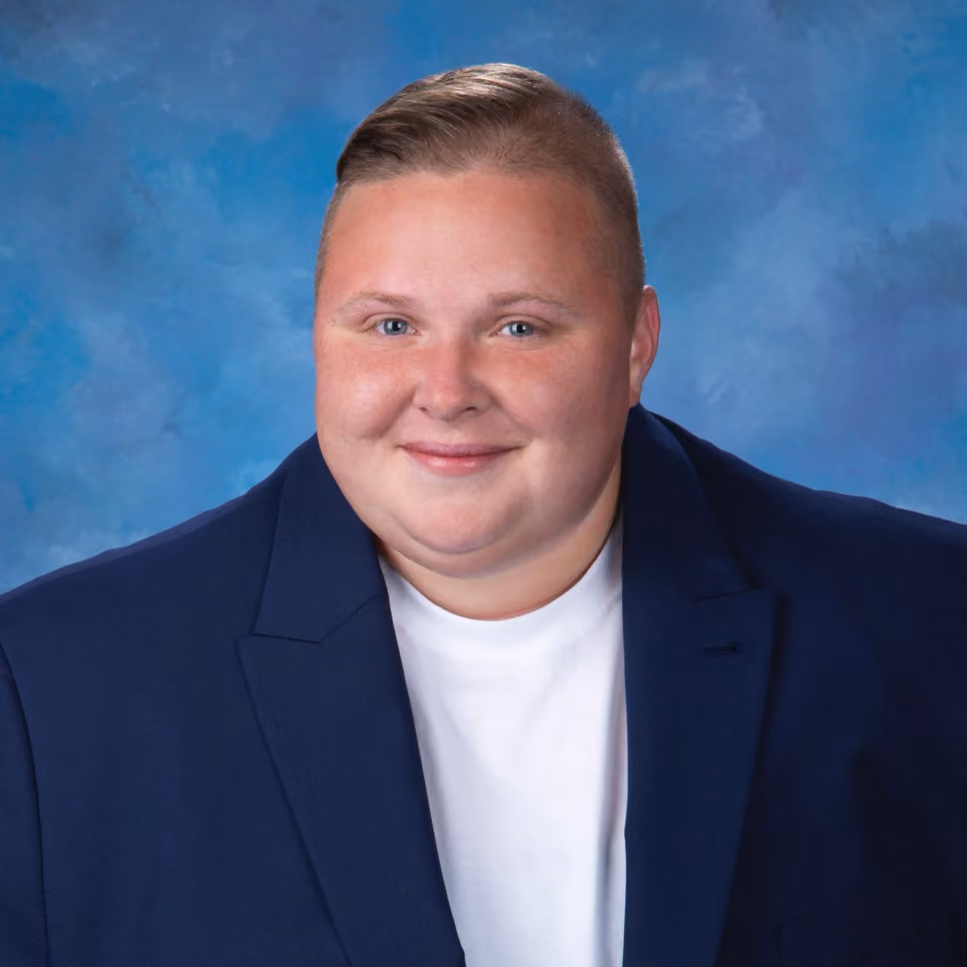 A young person with short, styled blond hair wearing a navy blue blazer over a white t-shirt, smiling in front of a blue studio backdrop.
