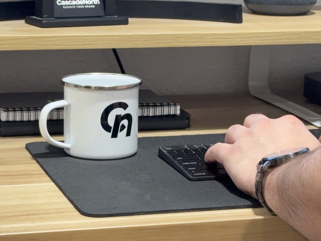 Hand wearing a wristwatch typing on a black keyboard next to a white metal mug with black 'Cn' logo on a wooden desk.