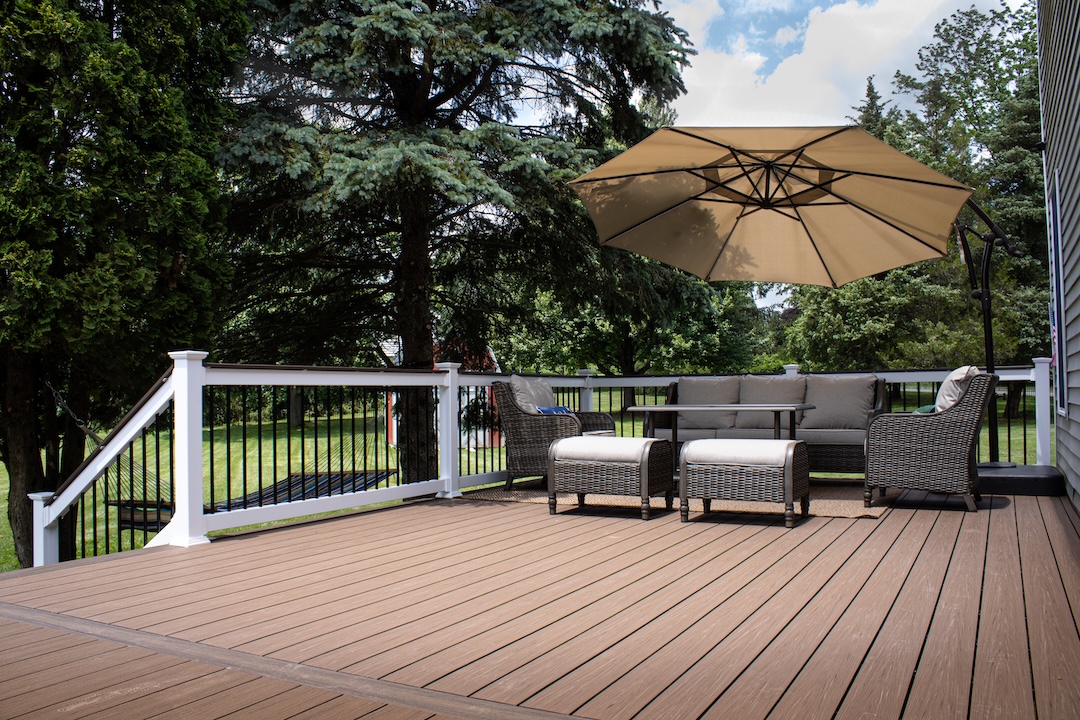 Composite deck with brown boards, white railings, wicker seating, and a tan umbrella in a wooded backyard.