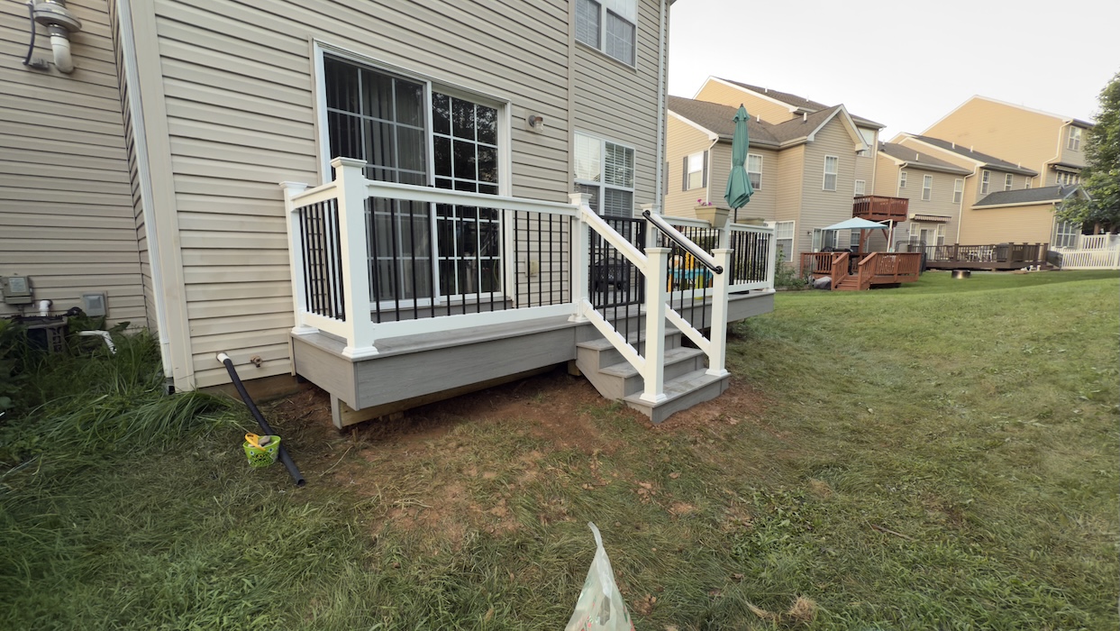 Small composite deck installation with white railing and black balusters attached to a suburban home, featuring gray decking and backyard steps.