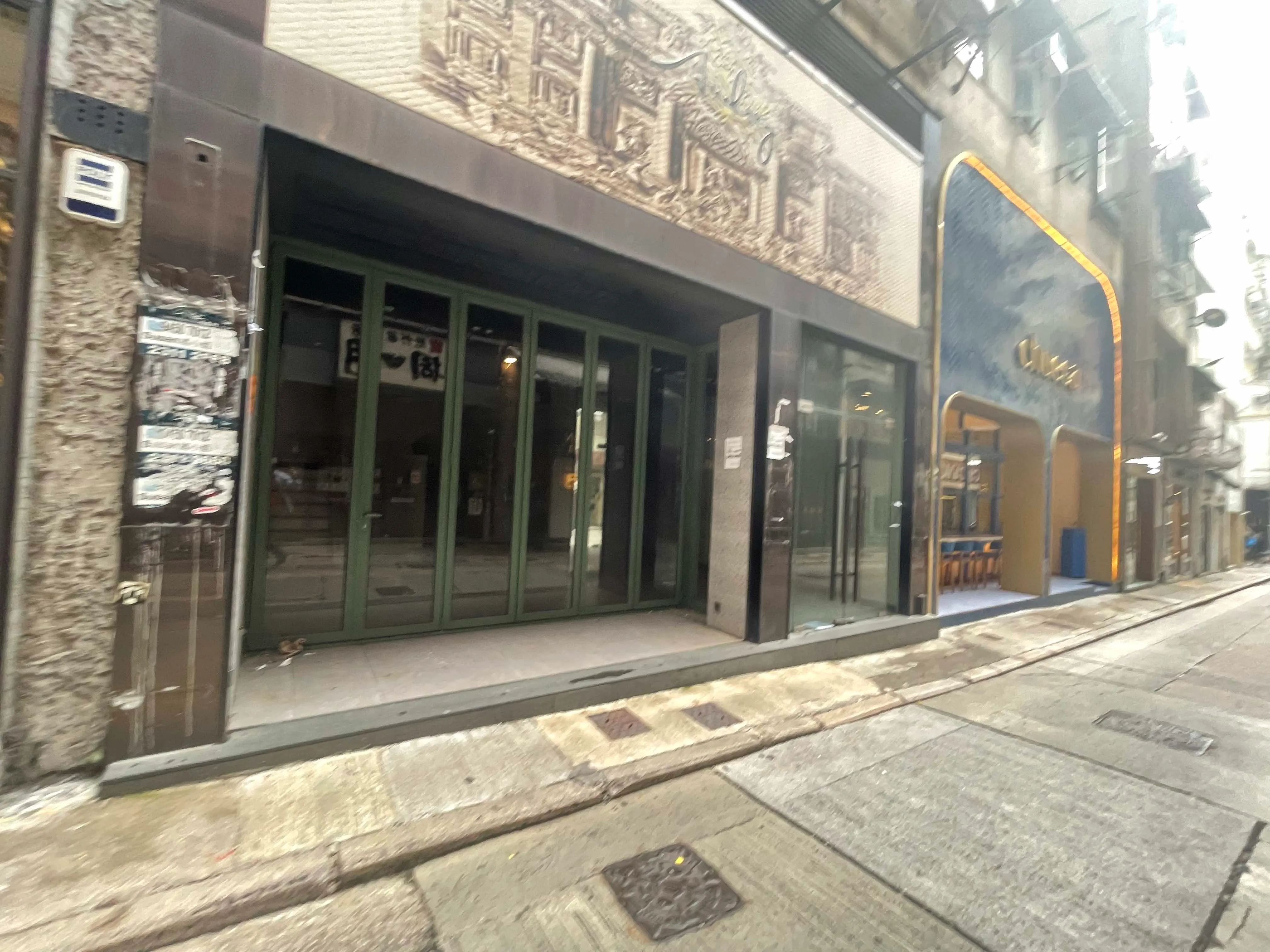 A blurry street view of Ref#2992 shows a closed storefront with glass doors and mural above, ideal for renting as shop space, situated next to a business with blue and gold signage; the sidewalk and street are empty.
