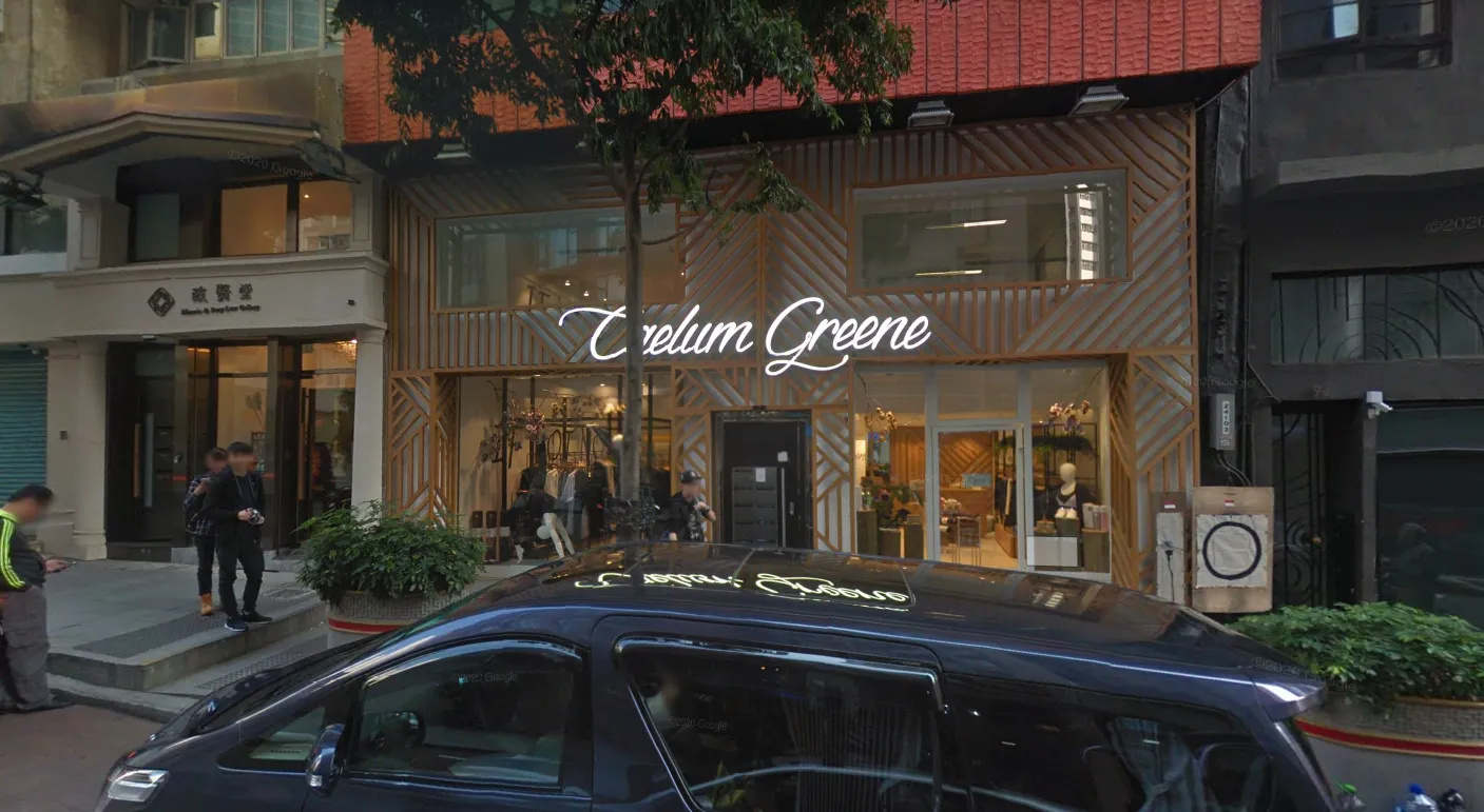 Ref#3005: A storefront with "Trelum Greene" in white script above large windows showcasing plants and decorideal for renting shop space. A black car is parked outside, people stand by the entrance, and trees and shops are nearby.