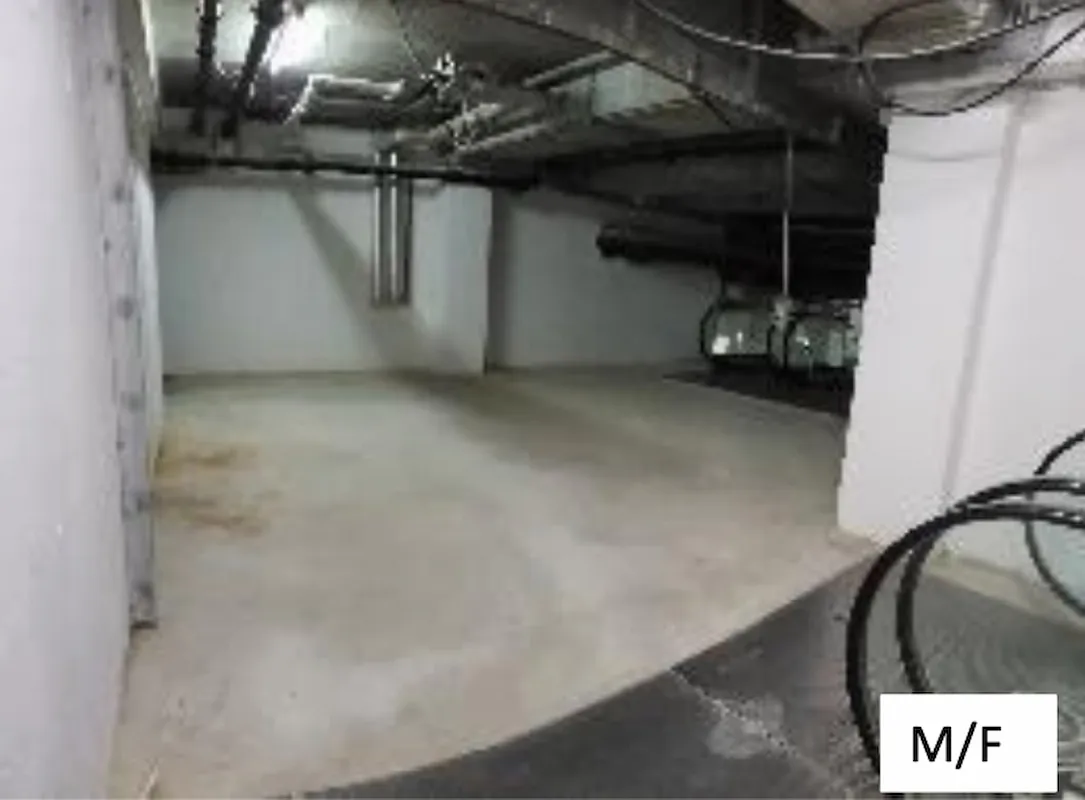 Ref#3011: A mostly empty underground parking garage with concrete floors, white walls, visible ceiling pipes, and a few cars on the far righterfect for office or shop rental. "M/F" in a white box is at the bottom right corner.