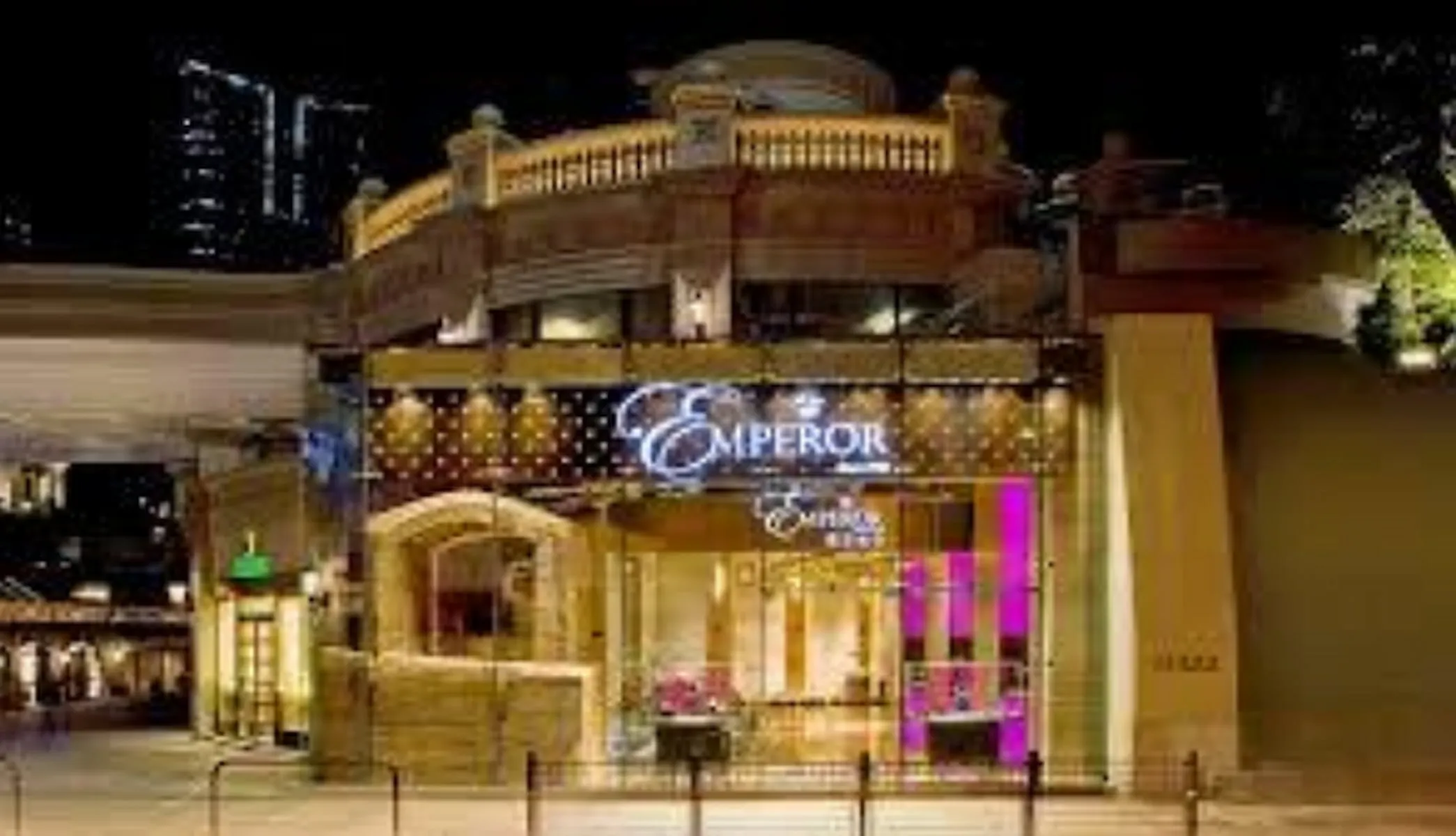 Exterior view of Ref#3011 at night, brightly lit with gold and purple lights. A large sign displays the name above the entrance, with urban buildings nearby offering shop and restaurant rental opportunities.