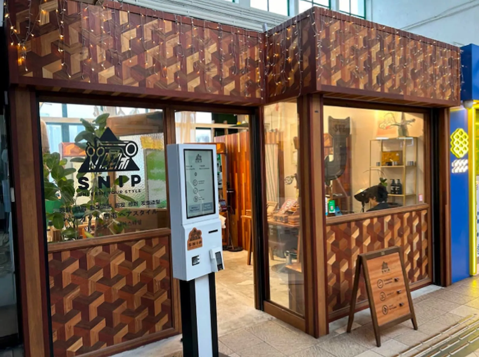 Ref#3018 features a compact shop with a geometric wood-patterned exterior, large windows, standing wooden sign, white kiosk outside, visible wooden goods and plants inside, plus string lights above the entranceideal for rental.